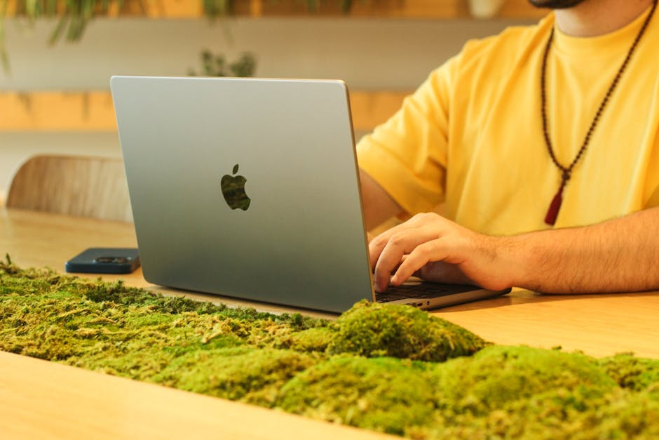 Man in yellow shirt working on laptop at a moss-decorated wooden table indoors.
