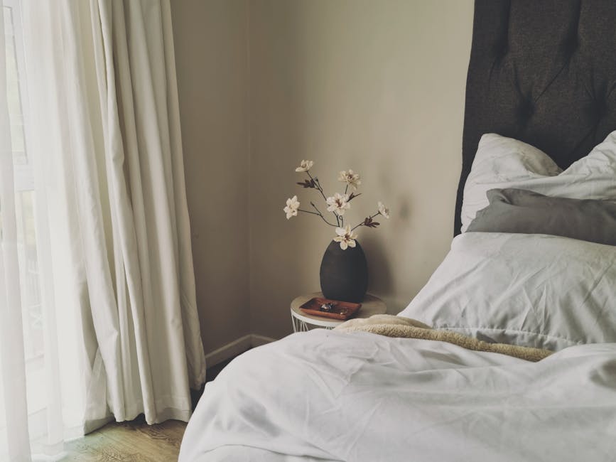 A serene and minimalist bedroom interior featuring white flowers in a vase, natural light, and a cozy bedding setup.