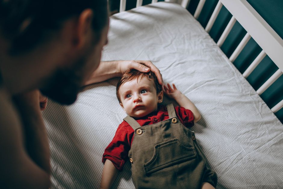 From above of unrecognizable bearded father stroking cute little boy in casual clothes lying in white crib in bedroom at home