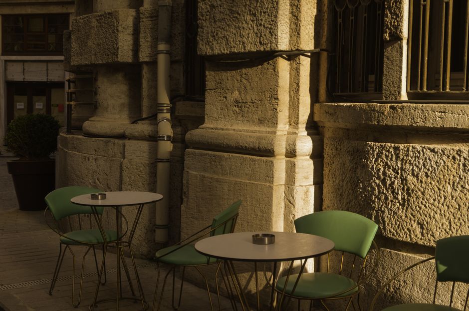 Outdoor café with green chairs and tables against an urban stone wall in warm sunset light.