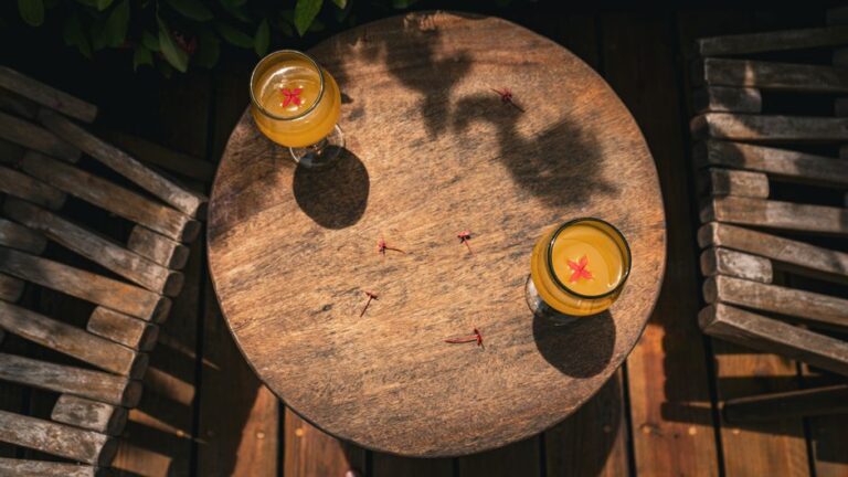 Overhead shot of two tropical drinks on a rustic wooden table outdoors, adorned with flowers.