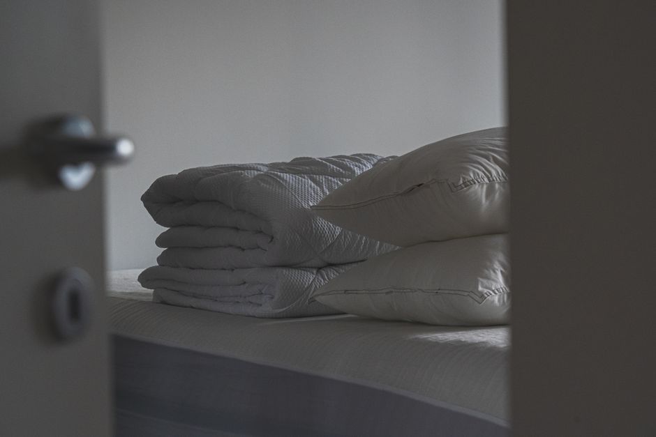 A serene bedroom scene featuring stacked pillows and folded blankets on a bed, viewed through an open door.