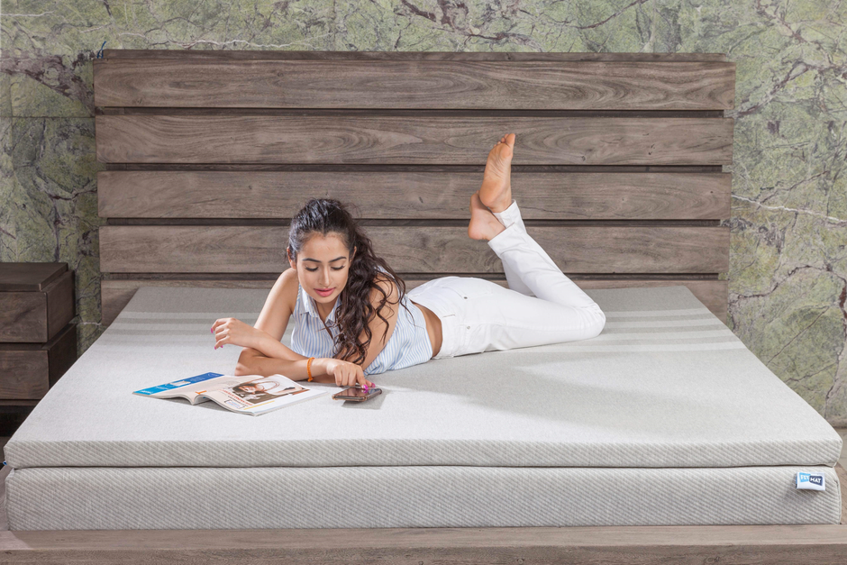 A young woman relaxing on a bed with a magazine and cellphone, set against a stylish wooden headboard.