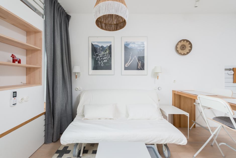 Cozy sofa bed with cushions placed in modern light studio apartment wooden table and shelves