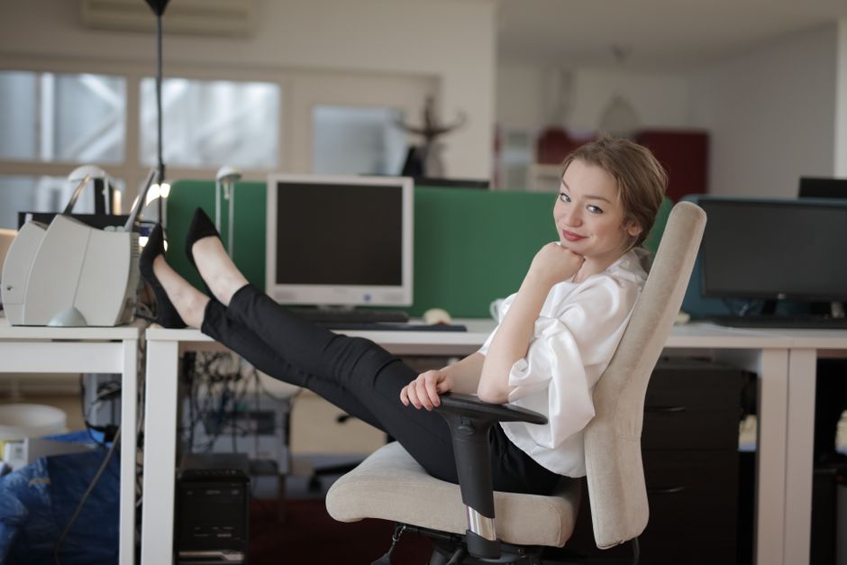Side view of cheerful female worker in formal style relaxing on office chair during break while sitting with feet on table and looking at camera