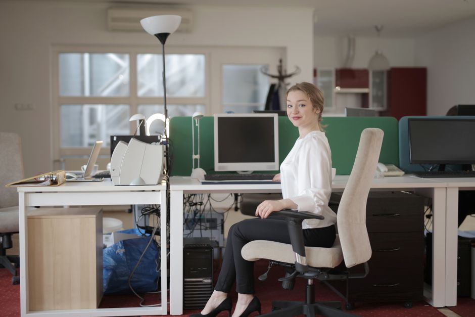Side view of glad female worker in classy outfit sitting at table on office chair during work in modern workplace and looking at camera