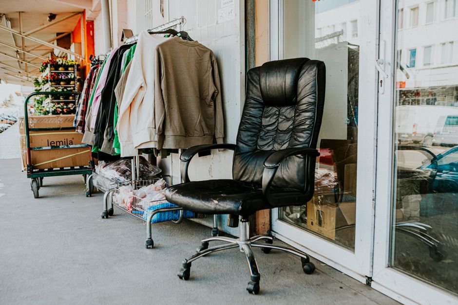 Worn black office chair outside a secondhand store with clothes and boxes nearby.