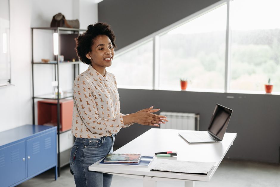 Young African American woman speaking at a desk with a laptop in a bright office.