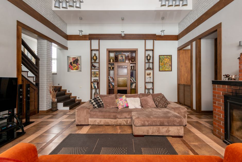 Interior of modern apartment with comfortable sofa and decorative elements on wooden shelves at wall in room with staircase and brick fireplace