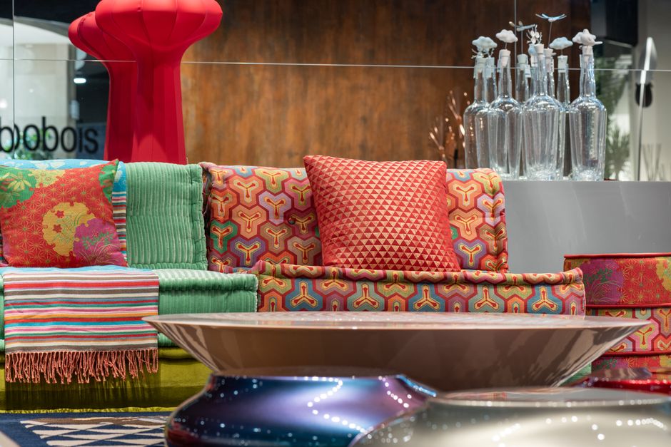 Multicolored comfortable sofa with bright cushions and vivid textile placed near modern round table in contemporary furniture store with glass wall and bottles